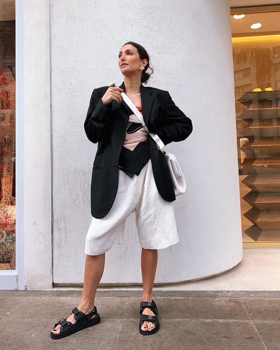 Mulher branca e jovem, de cabelo preto liso, posa para foto na rua, em frente a um muro branco. Ela usa um lenço de cedo amarrado como top, uma bermuda branca, um blazer preto e uma sandália rasteira preta. - Metrópoles