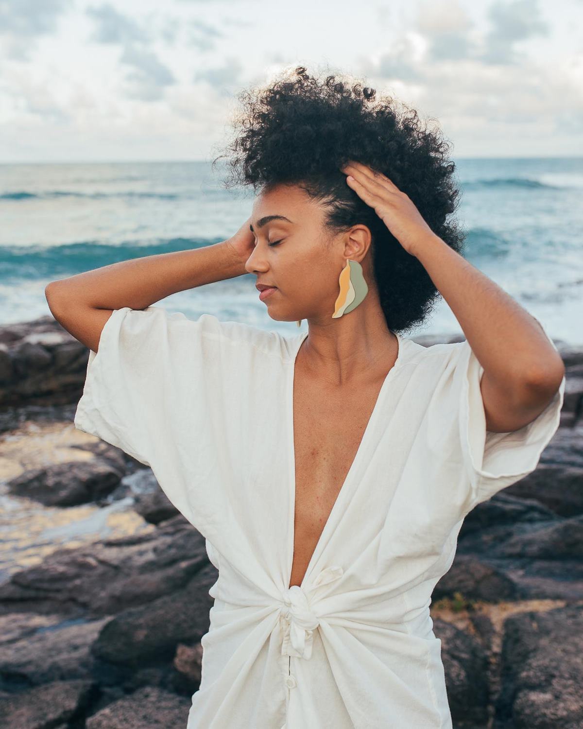 Mulher jovem e negra, de cabelo curto cacheado, posa para foto na beira da praia. Ela usa um vestido branco de linho, com um nó na frente, e um brinco grande nas cores amarela e verde. - Metrópoles