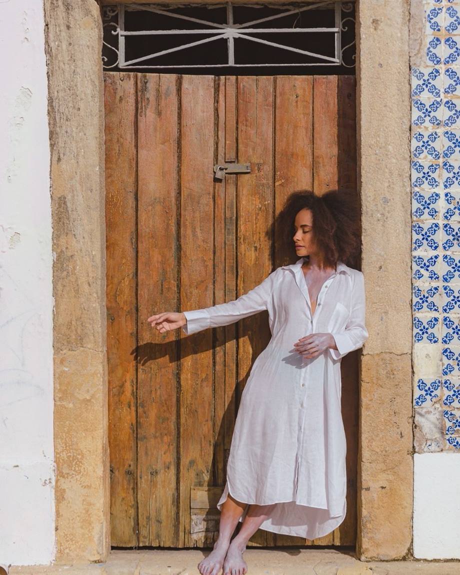 Mulher jovem e negra, de cabelo cacheado longo, posa para foto em frente a uma porta de madeira. Ela está descança e usa apenas uma chemise, que é um vestido que parece uma camisa de botão grande, na cor branca. - Metrópoles