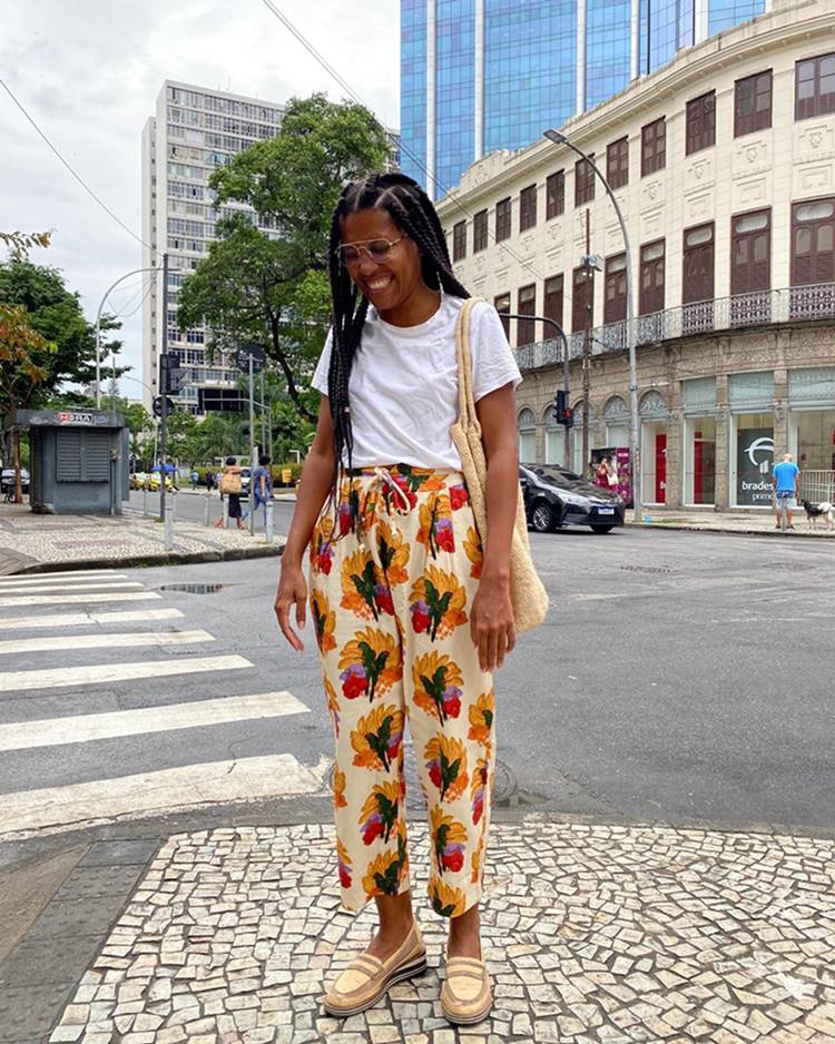 Mulher negra e jovem, de cabelo trançado longo, posa para foto nas ruas do centro do Rio de Janeiro. Ela usa óculos de grau e veste uma camiseta branca, uma calça com flores estampadas, um mocassim branco e uma bolsa de palha. - Metrópoles