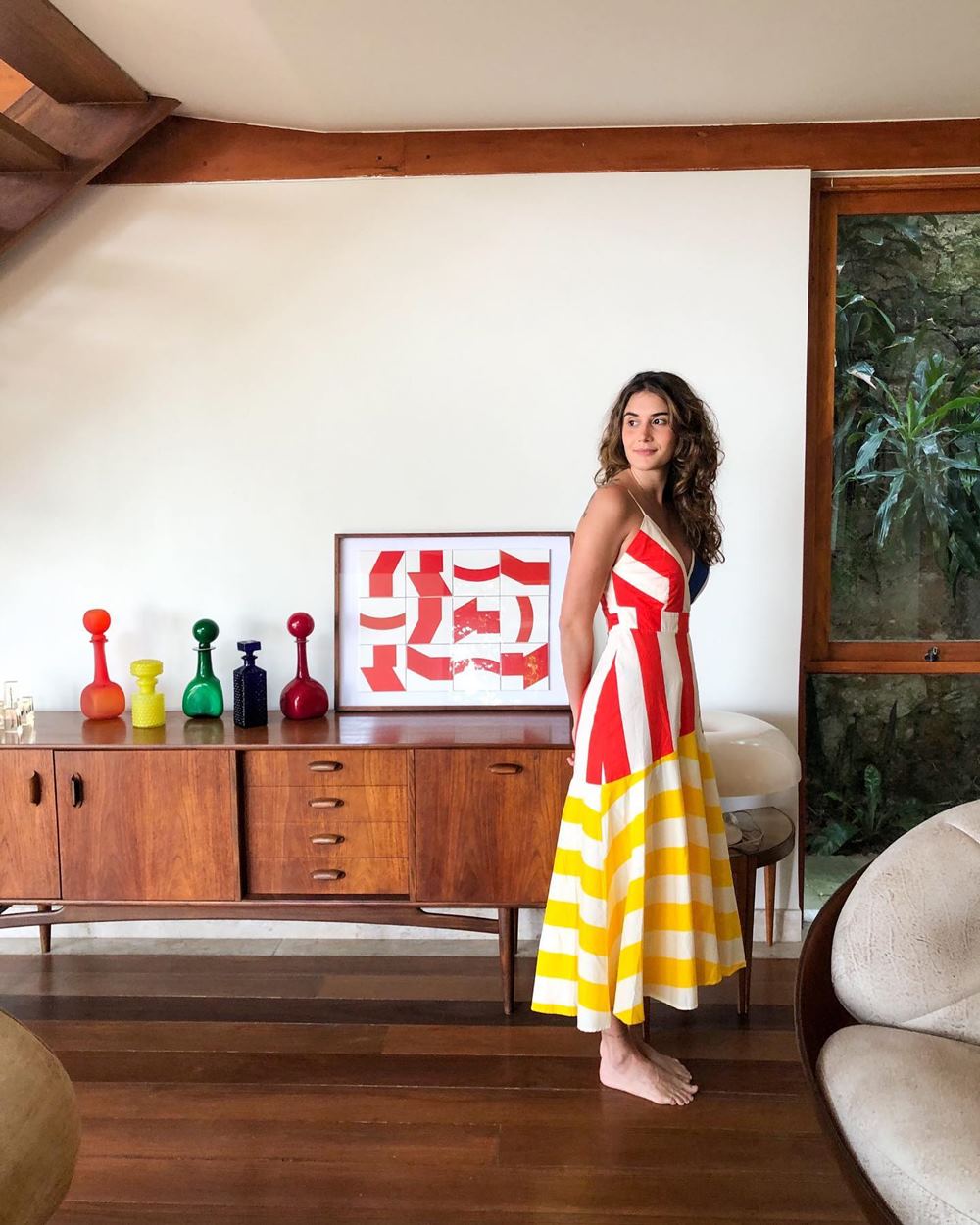 Mulher branca e jovem, de cabelo ondulado castanho, posa para foto na sala de casa, ao lado de uma cômoda de madeira. Ela usa um vestido listrado com a parte de cima vermelha e branca e, a de baixo, amarela e branca. A peça é da loja Farm. - Metrópoles