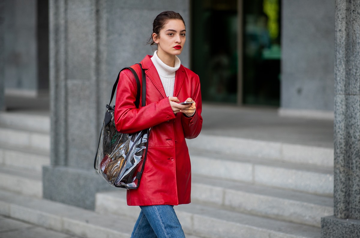 Mulher branca e jovem, de cabelo liso castanho amarrado em um coque, andando pela rua. Ela usa um casaco branco de tricô com gola alta, um casaco vermelho de couro, uma calça jeans e uma bolsa preta larga. Os lábios estão maquiados com batom vermelho. - Metrópoles