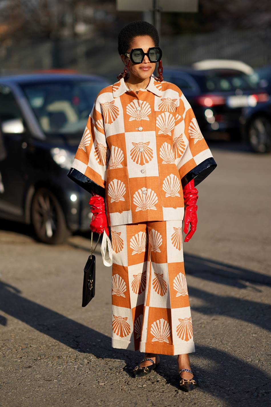 Mulher jovem e negra, de cabelo cacheado curto, posa para foto em um estacionamento de carros. Ela usa um conjunto de camisa e calça, com estampa branca e laranja, uma luva vermelha e uma sapatilha preta. - Metrópoles