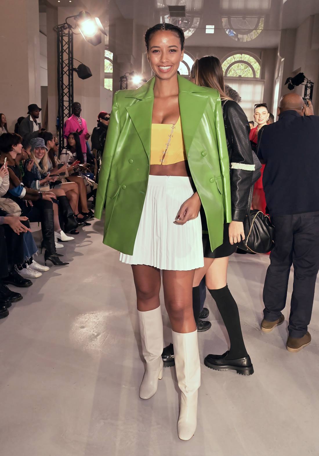 Mulher negra e jovem, com cabelo cacheado amarrado, posa para foto em um desfile. Ela usa um top amarelo, uma saia branca plissada, uma jaqueta verde couro e uma bota branca. - Metrópoles