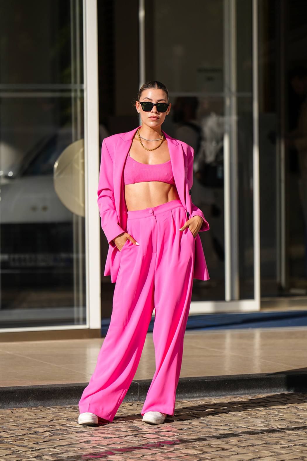 Mulher branca e jovem, de cabelo liso castanho amarrado em um coque, posa para foto em frente a um prédio comercial. Ela usa um top rosa e um conjunto de blazer e calça social, tudo também rosa. - Metrópoles