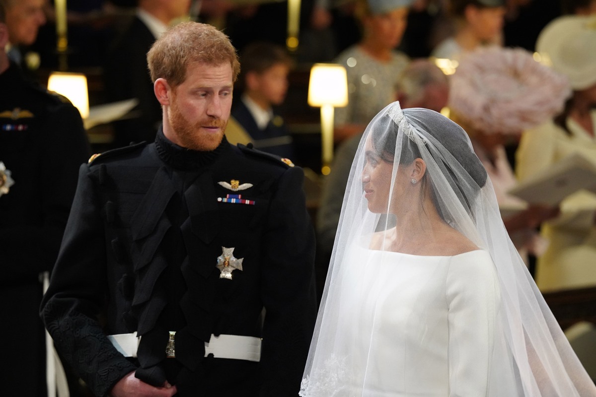 O princípe Harry, um homem branco e jovem, de cabelo liso ruivo, ao lado da esposa, a atriz Meghan Markle. Ela é uma mulher jovem negra, de cabelo castanho longo. Ele está com uma roupa preta militar e ela com um vestido branco de noiva e véu cobrindo o rosto. O registro é do casamento de Harry e Meghan. - Metrópoles