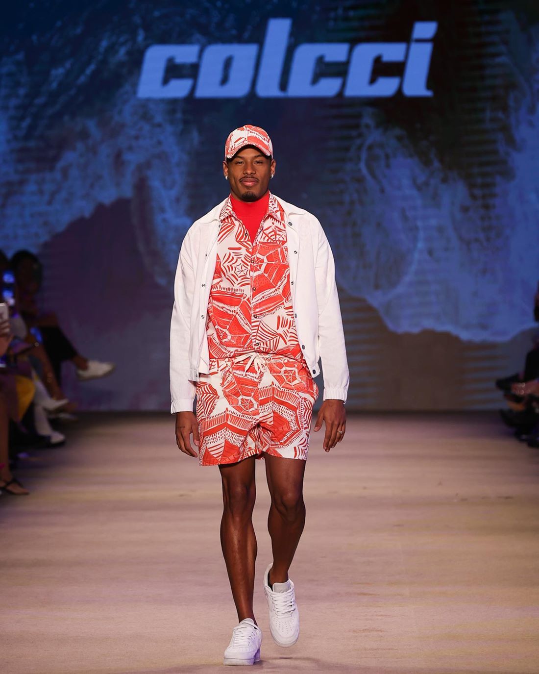 O ex-BBB e atleta Paulo André, um homem jovem e negro, de cabelo cacheado curto, desfila na passarela da marca Colcci no São Paulo Fashion Week. Ele usa um conjunto de camisa de botão e shorts, ambos com uma estampa de folha vermelha e branca, um casaco branco por cima e tênis também branco. - Metrópoles