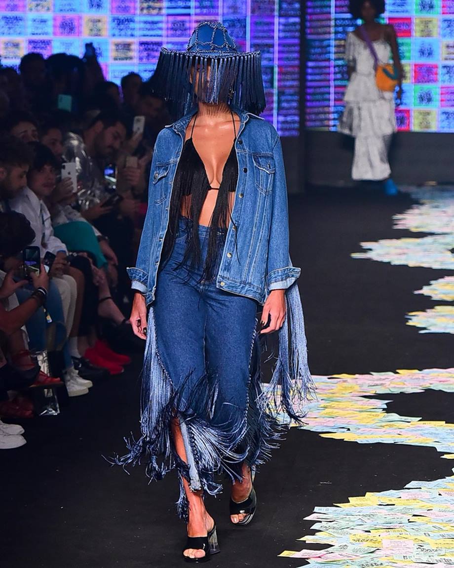 A ex-BBB e modelo Mari Gonzalez, uma mulher jovem e branca, de cabelo liso longo, em desfile no São Paulo Fashion Week. Ela usa um top preto de franjas, uma calça e uma jaqueta, ambos jeans, e um chapéu, também com franjas e também jeans. - Metrópoles