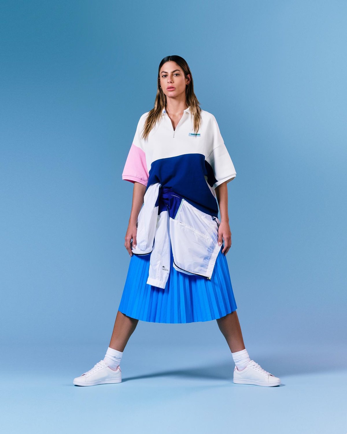 A ex-BBB e modelo Mari Gonzalez, uma mulher jovem e branca, de cabelo liso longo, posa para foto em um fundo liso azul. A foto é de uma campanha publicitária da marca Lacoste. Ela usa uma camisa polo branca, uma saia plissada azul, tênis branco e, amarrada à cintura, uma jaqueta estilo esportiva azul escura. - Metrópoles