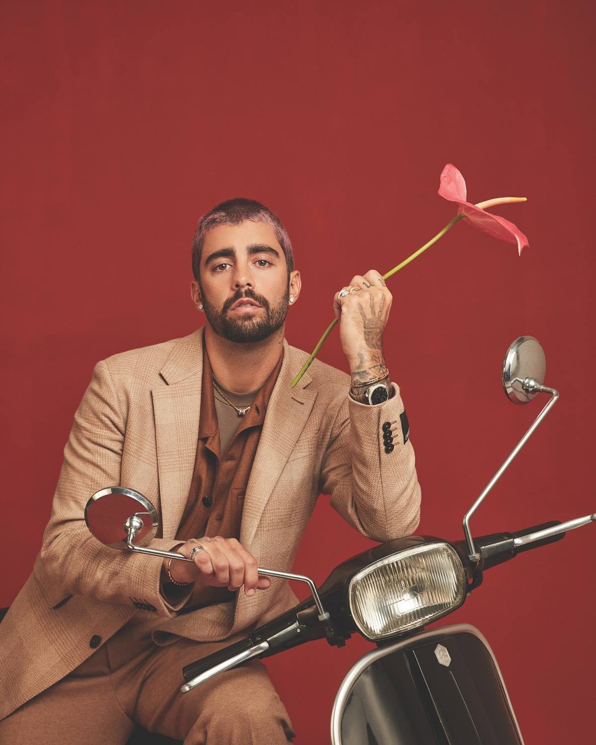 O ex-BBB e surfista Pedro Scooby, um homem branco e jovem, de cabelo e barba castanhos, posa para foto em um fundo vermelho. Ele usa uma camisa marrom, um blazer bege e segura uma flor com uma das mãos. Está em cima de uma moto. - Metrópoles