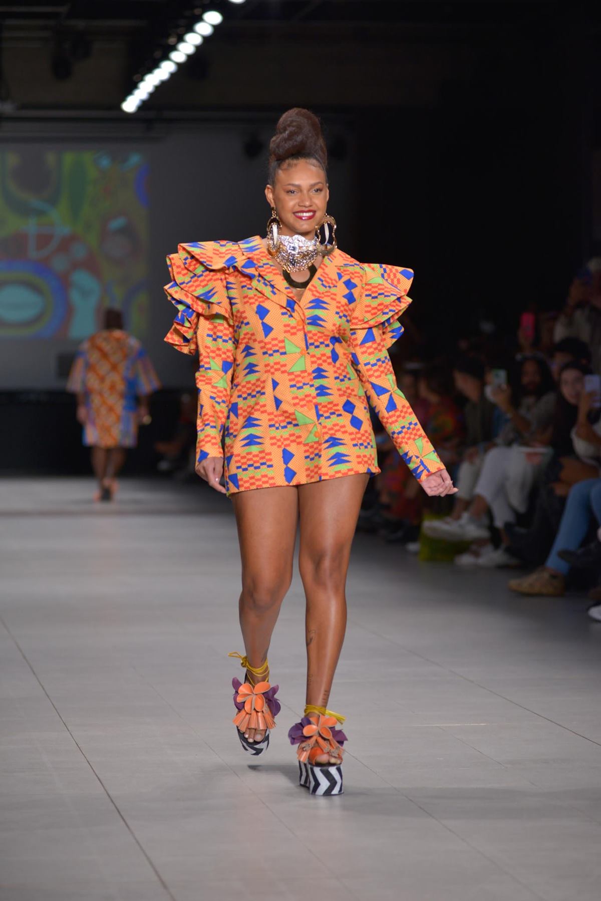 A ex-BBB Jessilane, uma mulher jovem e negra, de cabelo cacheado amarrado em um coque, cruza a passarela do São Paulo Fashion Week em desfile da marca Meninos Rei. Ela usa um vestido curto com mangas bufantes e babados nos ombros. - Metrópoles