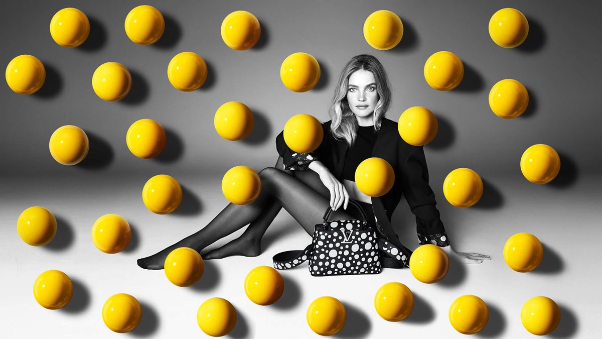 A modelo Natalia Vodianova, uma mulher branca e jovem, com cabelo liso loiro, posa para campanha da marca Louis Vuitton. Ela usa um top curto, um shorts e um blazer, tudo preto; além de uma bolsa de couro preta com bolas brancas. - Metrópoles