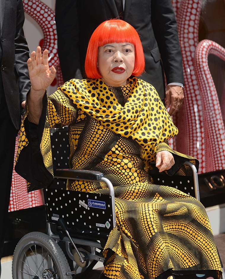 A artista plástica Yayoi Kusama, uma senhora japonesa idosa, com uma peruca laranja em corte Chanel, em um evento da Louis Vuitton. Ela usa um vestido longo amarelo com bolas pretas e está sentada em uma cadeira de roda. - Metrópoles