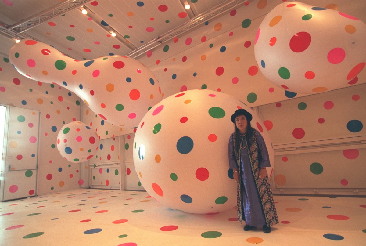 A artista plástica Yayoi Kusama, uma mulher de meia idade japonesa, em uma exposição de arte com suas obras. Ela usa um vestido roxo longo e um quimono cinza longo, além de um chapéu preto. A foto é de 2001. - Metrópoles