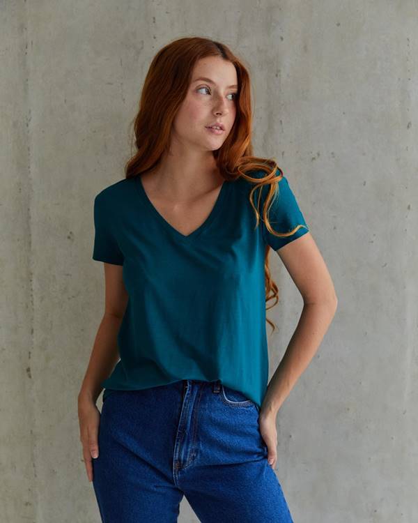 Modelo branca e ruiva e parede de fundo de cimento queimado usa camiseta azul e calça jeans - Metrópoles 