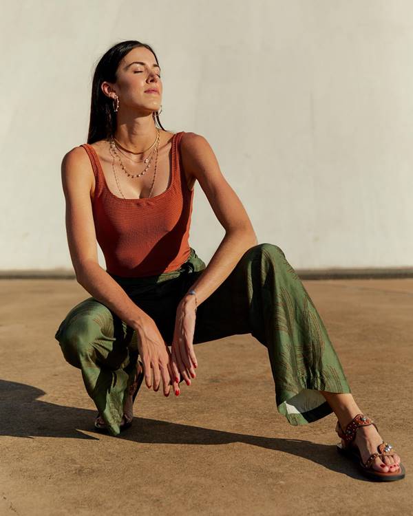 Modelo branca em posição agachada usa calça verde escura e top marrom. O look é completado com acessórios - Metrópoles 