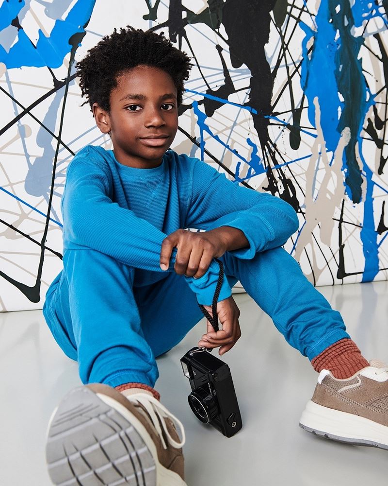 Menino jovem e negro, de cabelo cacheado curto, posa para foto em um fundo branco com pinceladas de tinta. A foto é de campanha da marca Hering Kids. Ele usa um conjunto de casaco e calça, ambos de moletom azul, e um tênis cinza com detalhes brancos. - Metrópoles