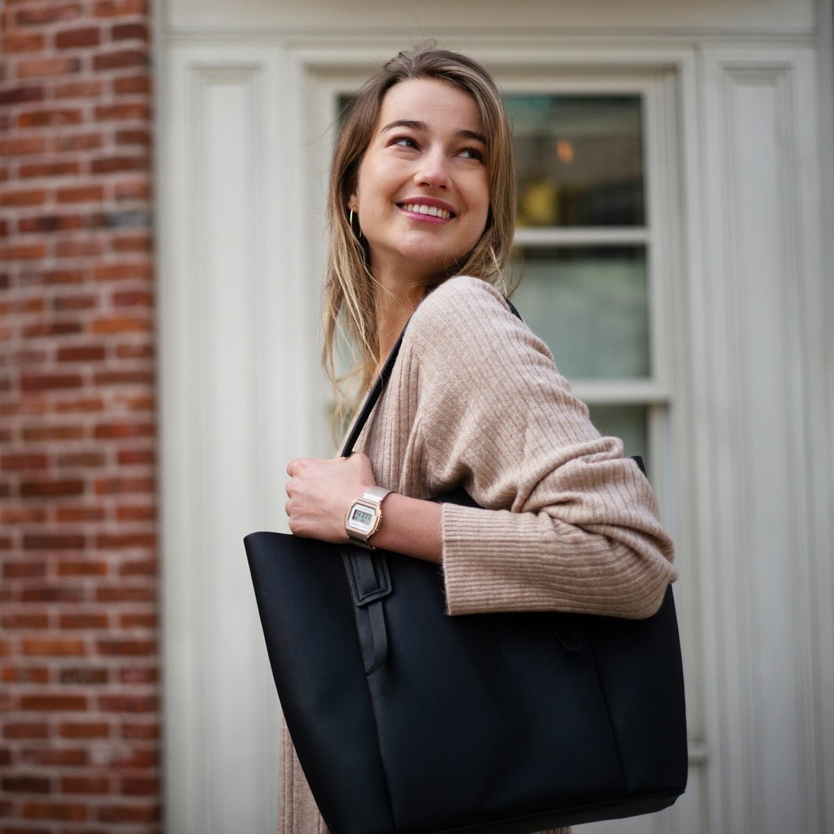 Mulher branca e jovem, de cabelo liso loiro, posa para foto. Ela usa um casaco bege, uma bolsa de couro preta e um relógio dourado da marca Casio. - Metrópoles