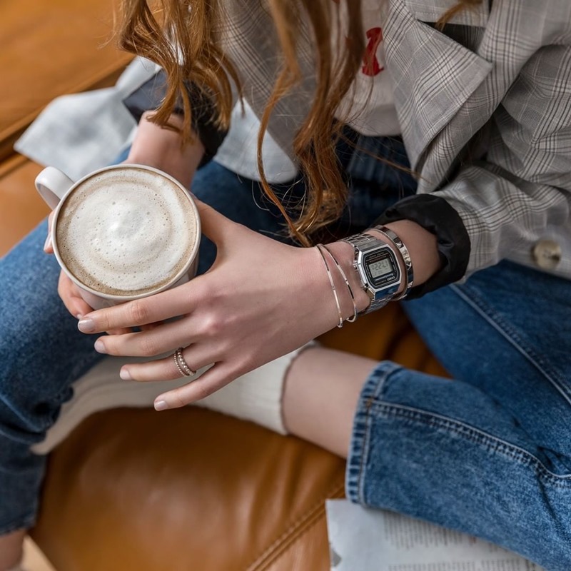 Mulher branca e jovem, de cabelo liso castanho, posa para foto. Ela usa um casaco cinza, calça jeans, meias brancas e um relógio dourado da marca Casio. - Metrópoles