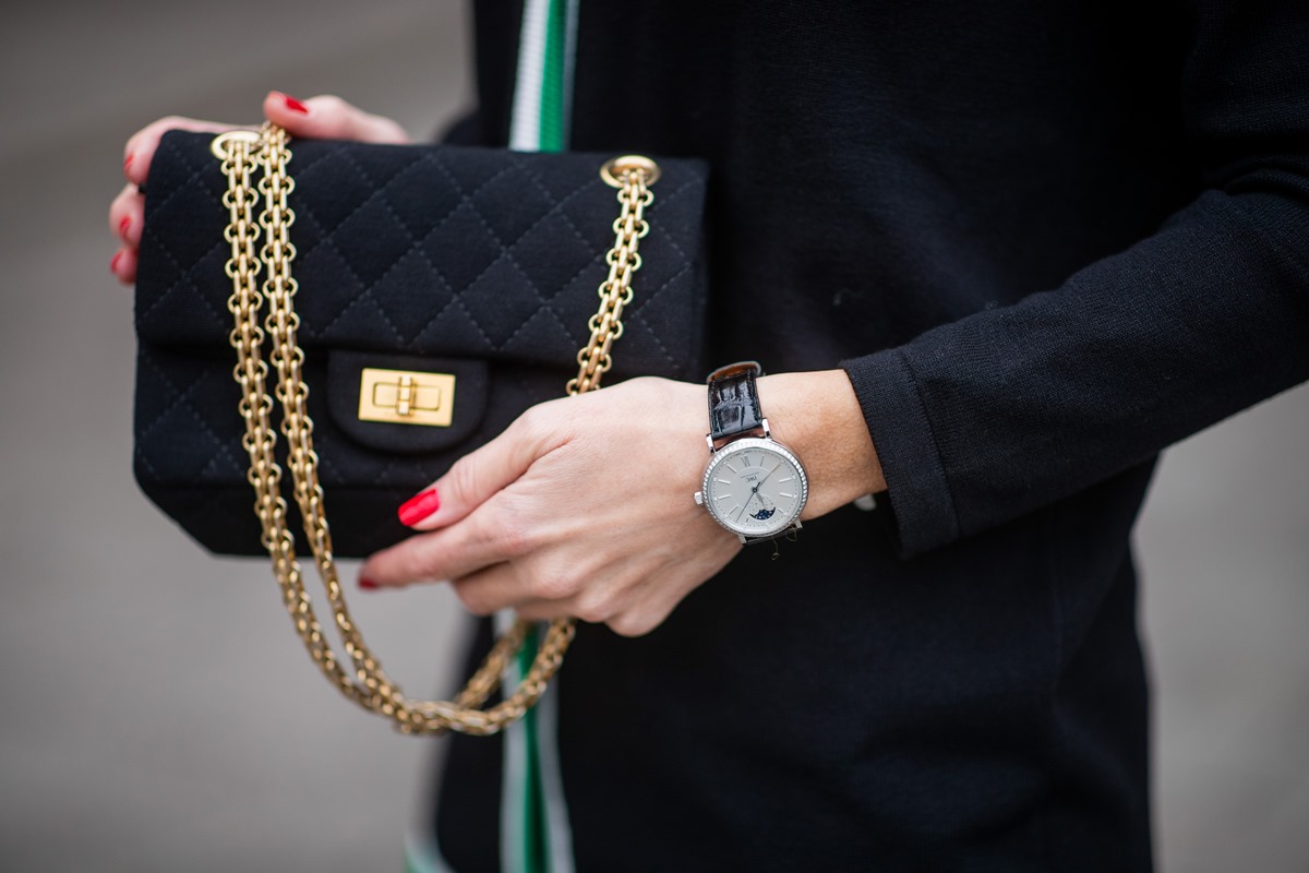 Mulher branca e jovem segura uma bolsa preta da marca Chanel. A peça é preta e de camaruça e com correntes e os fechos dourados. A mulher usa um blazer preto e um relógio preto, de couro, com o ferragens pratas. - Metrópoles