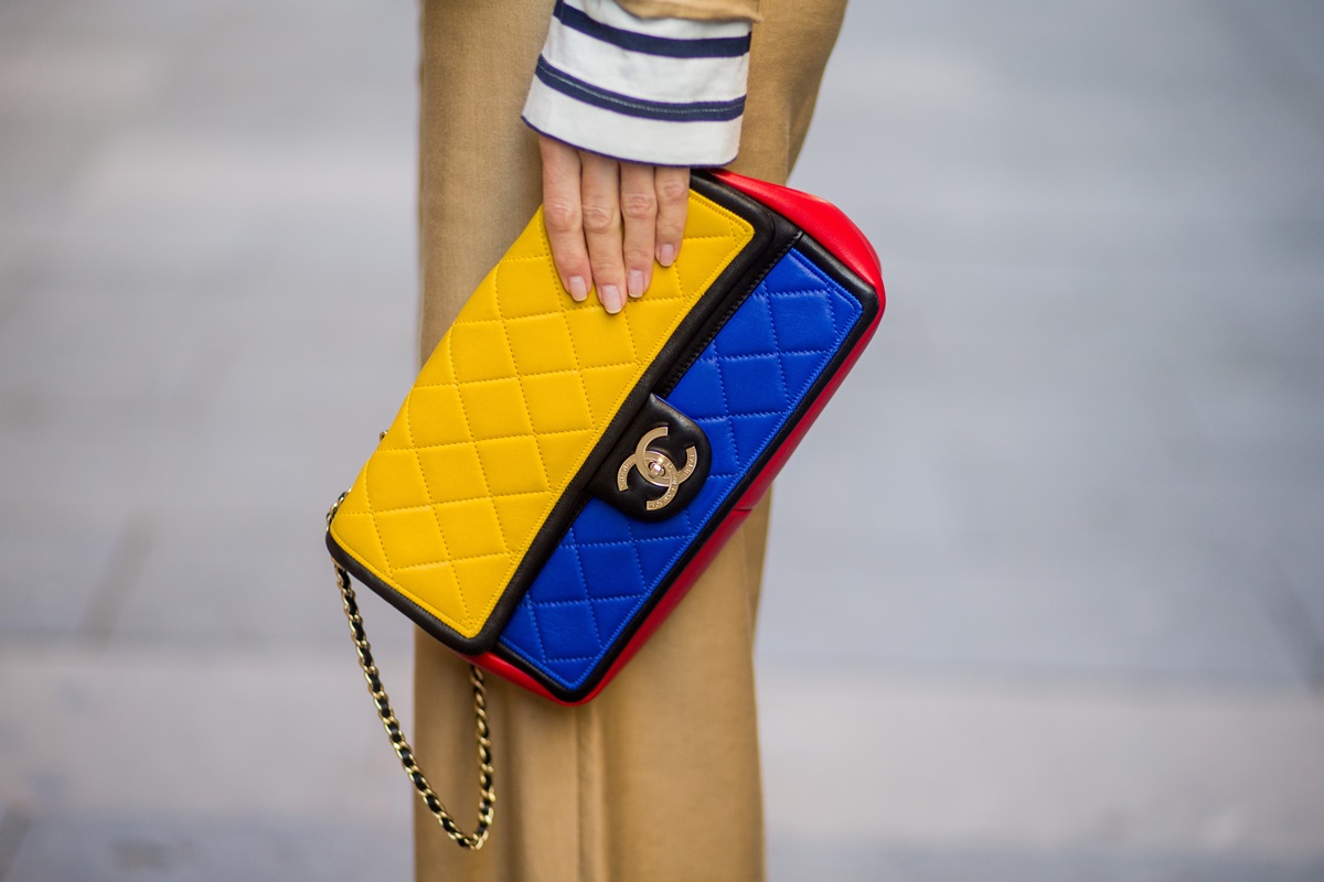 Mulher branca e jovem segurando uma bolsa modelo 2.55 da marca Chanel. Na foto não é possível ver seu rosto, apenas suas mãos e suas pernas. A bolsa é nas cores amarela, azul e vermelha com detalhes pretos. Ela usa uma blusa branca com listra azuis e uma calça marrom. - Metrópoles