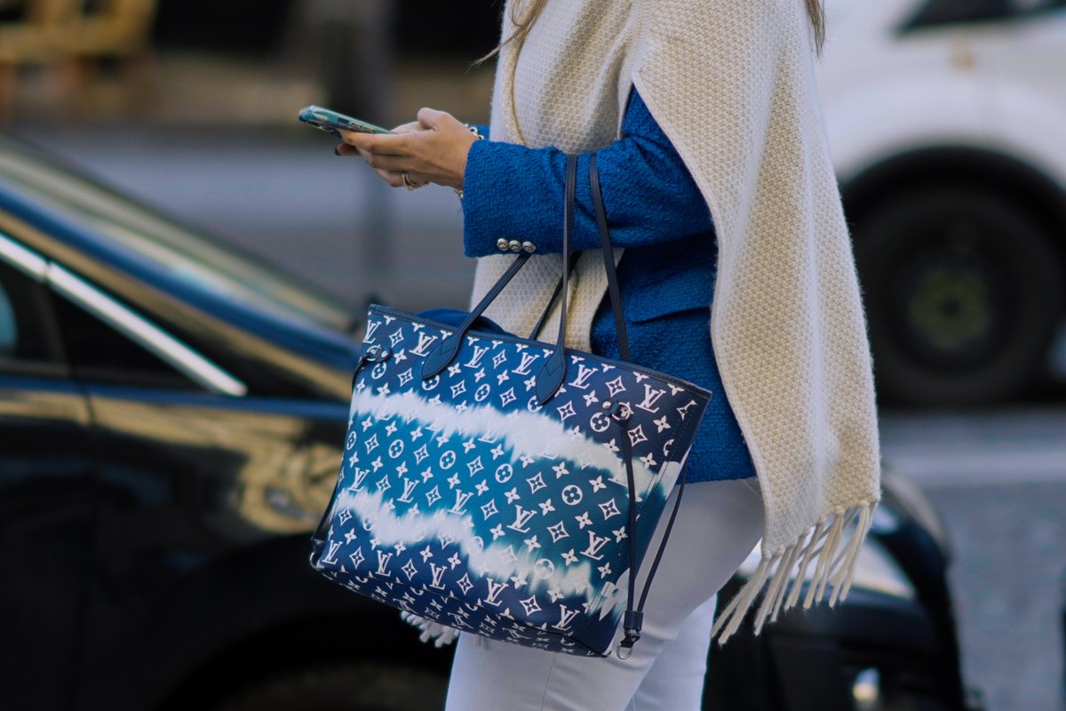 Bolsa modelo Neverfull, da marca Louis Vuitton. A peça é azul de couro com a logomarca da grife estampada. - Metrópoles