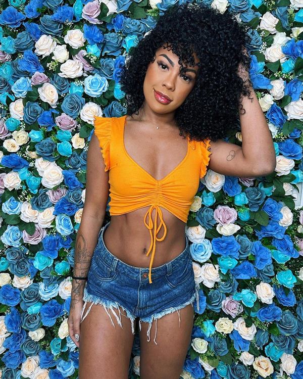 Mulher negra com shorts jeans e top amarelo. Ela está em frente a um fundo de flores - Metrópoles