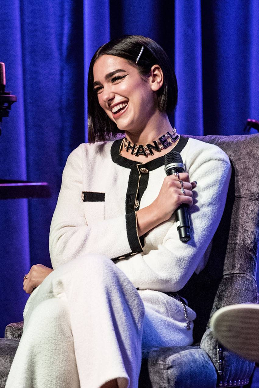 A cantora Dua Lipa, uma mulher jovem e branca, de cabelos curtos pretos e lisos, dando entrevista. Ela segura um microfone e usa um conjunto de blazer e calça de tweed. As peças são da marca Chanel. - Metrópoles