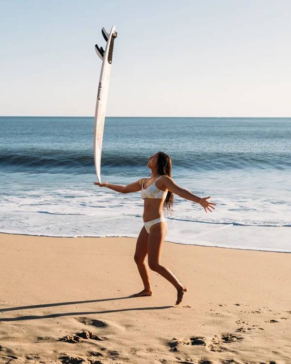 Sophia Medina com biquini branco ao lado de uma prancha de surfe na praia - Metrópoles