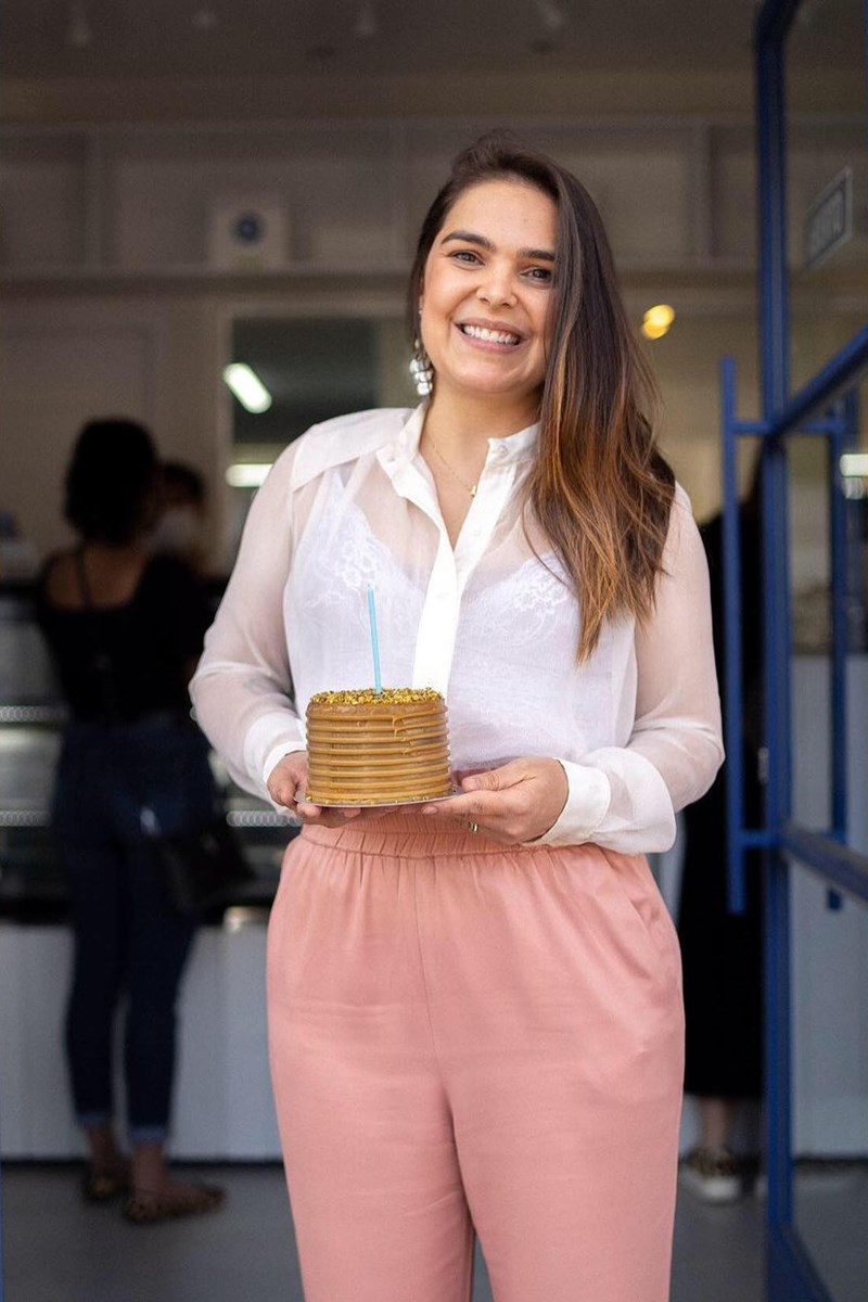 Foto colorida de mulher parda, com cabelo castanho claro, segurando um bolo em frente a uma porta - Metrópoles