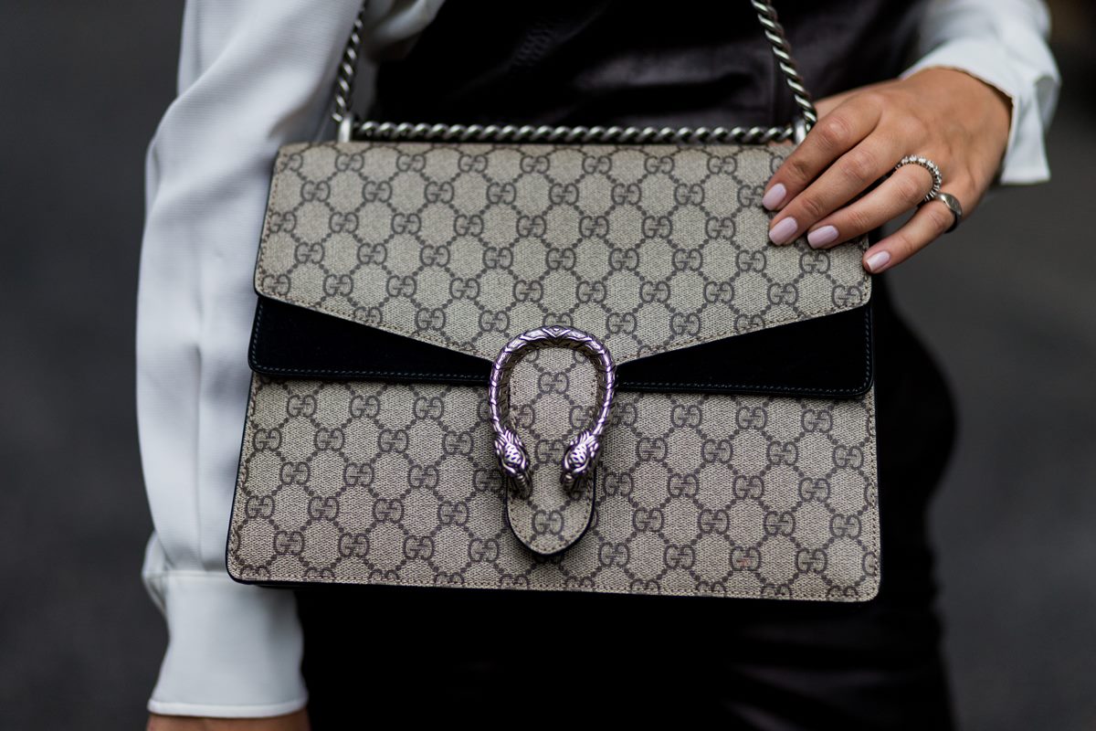 Mulher branca e jovem mostra sua bolsa da Gucci. O modelo da bolsa se chama Dionysus. É uma bolsa bege de lona com um fecho prata, em formato de cobra, e alça de corrente, também prata. - Metrópoles