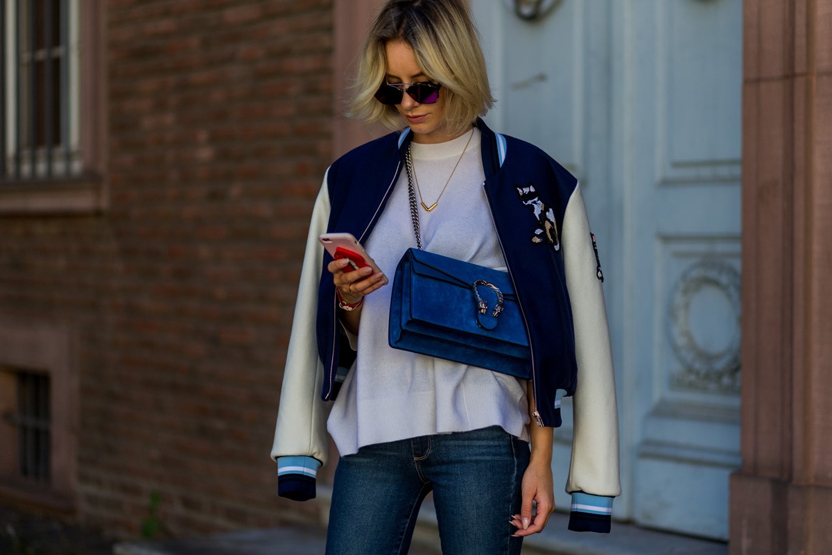 Mulher branca e jovem, de cabelo loiro liso, mexe no celular. Ela usa uma camiseta branca, uma calça jeans, uma jaqueta branca e azul estilo colegial, óculos escuros e a bolsa modelo Dionysus da Gucci. - Metrópoles