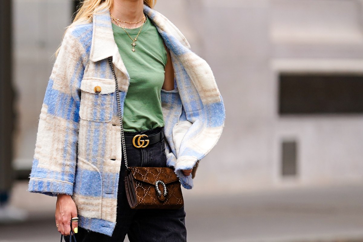 Mulher branca e jovem, de cabelo loiro liso, andando pela rua. Ela usa uma regata verde, calça jeans preta, um casaco xadrez branco e azul e uma bolsa Dionysus marrom, de camurça, da Gucci. - Metrópoles