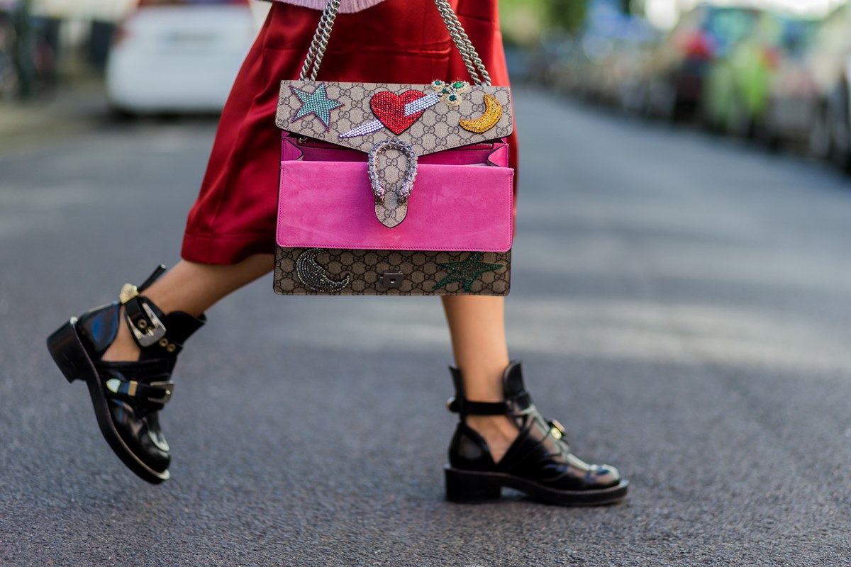 Mulher branca andando pela rua. Ela usa um vestido vermelho e uma bolsa de couro da marca Balenciaga. Segura, em uma das mãos, uma bolsa marrom de couro chmada Dionysus da marca Gucci. - Metrópoles