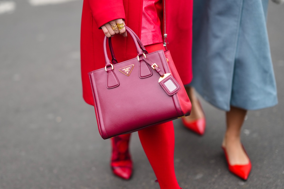 Mulher branca andando pela rua e segurando uma bolsa vermelha de couro, modelo Galleria, da marca Prada. Ela usa um look tudo vermelho: sapatos de salto, meia calça e casaco. - Metrópoles