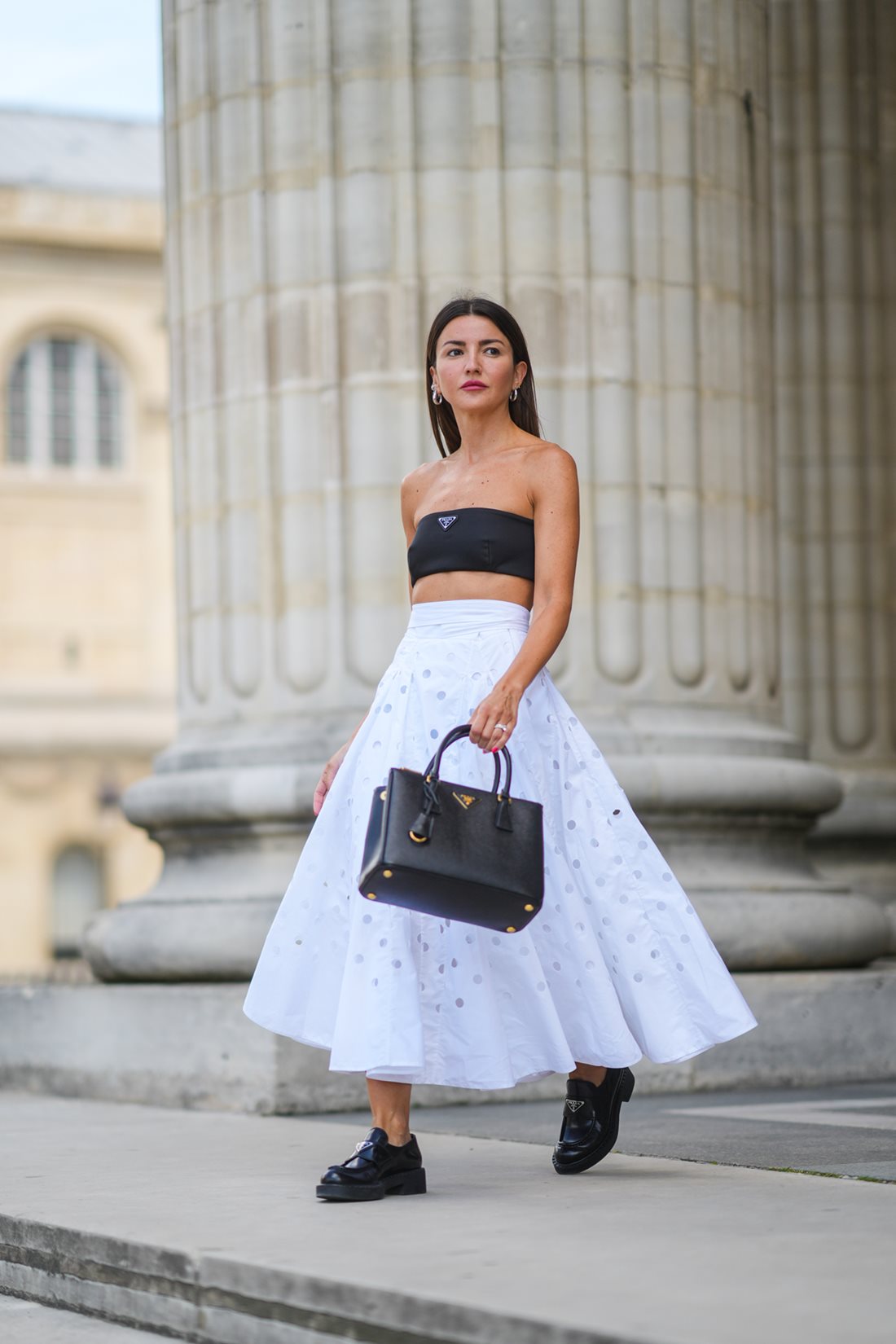 Mulher branca e jovem, de cabelo liso preto, andando pela rua e posando para foto. Ela usa um top faixa sem alça preto, uma saia rodada branca, sapatos mocassim preto e uma bolsa de couro chamada Galleria. Todas as peças são da grife Prada. - Metrópoles