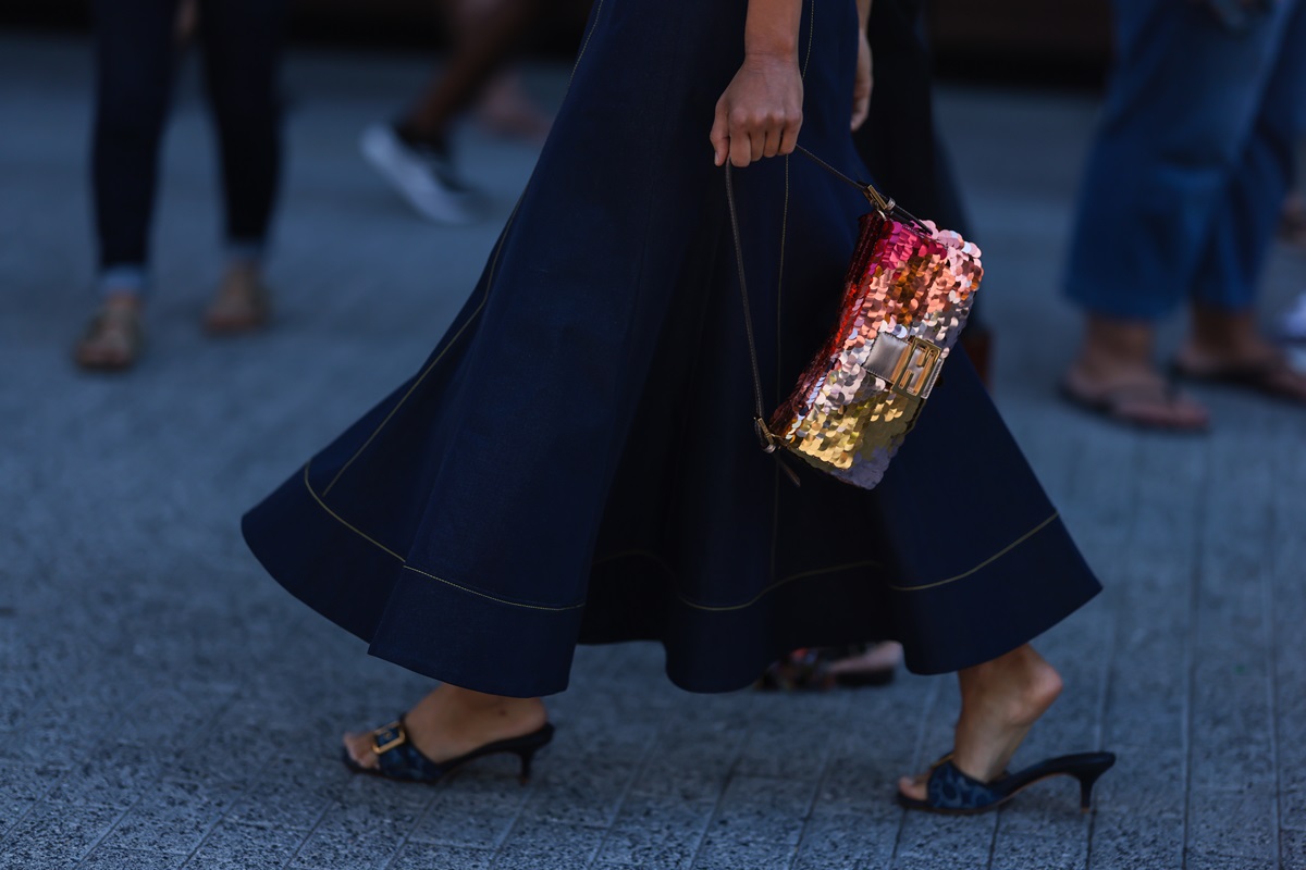 Mulher negra e jovem andando pela rua. Ela usa um vestido longo e jeans, um salto escuro e uma bolsa Baguette da marca Fendi. A peça é feita de paetês nas cores laranja, rosa e amarelo. - Metrópoles