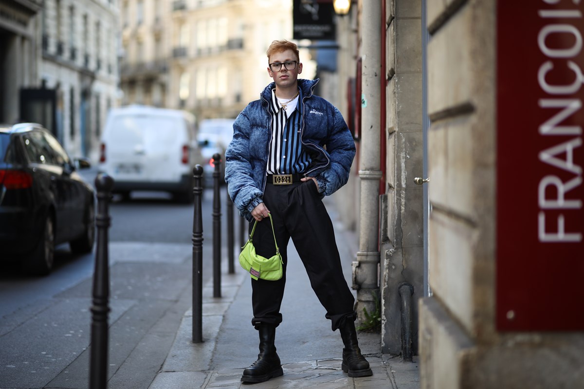 Homem jovem e branco, de cabelo ruivo liso, posa para foto nas ruas de Paris, na França. Ele usa uma camisa listrada de botão, calça preta, jaqueta puffer azul, bota de couro preta e uma bolsa Baguette verde limão da marca Fendi. - Metrópoles