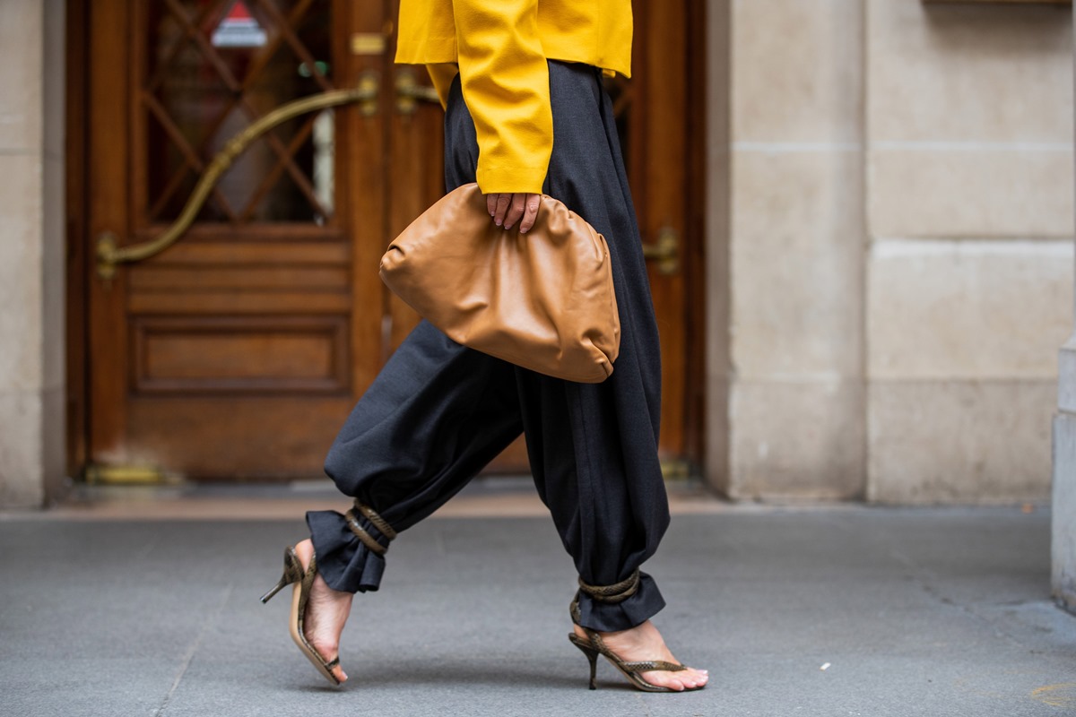 Mulher negra e jovem caminha pela rua. Ela usa uma calça preta folgada, um casaco amarelo, um sandália de salto marrom e uma bolsa, modelo The Pouch, da marca Bottega Veneta, na cor marrom. - Metrópoles