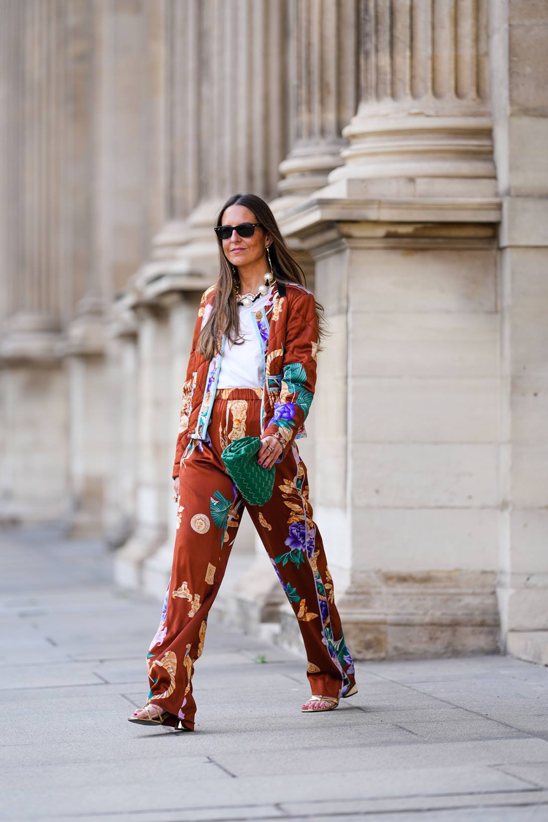 Mulher branca e de meia idade, com cabelo liso castanho e longo, andando pelas ruas de Paris, na França. Ela usa uma camiseta branca, um conjunto de casaco e calça marrom com estampas coloridas, uma bolsa verde de couro, modelo The Pouch, da marca Bottega Veneta, e óculos escuros Rayban. - Metrópoles