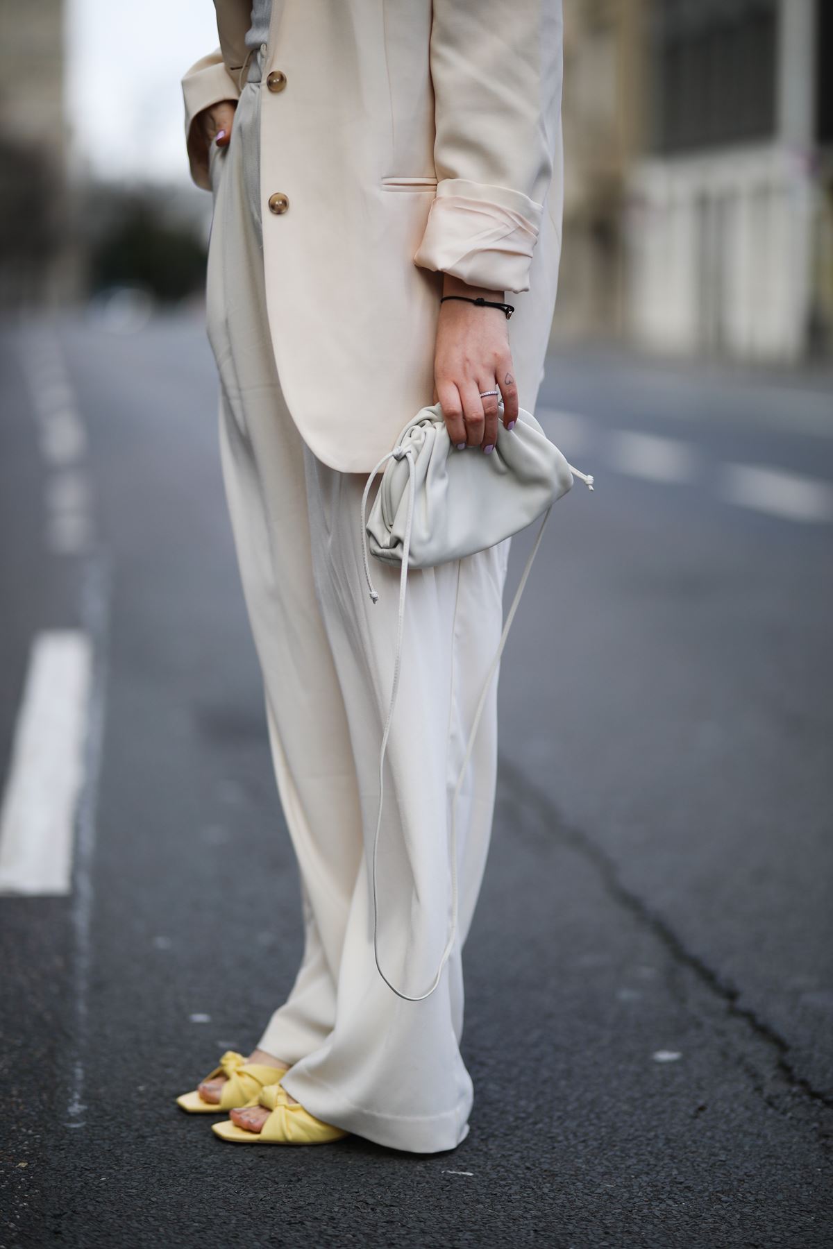 Foto das pernas de uma pessoa durante a Semana de Moda de Paris. A mulher, que parece ser branca e jovem, usa um conjunto de blazer e calça social, ambos na cor creme; uma sandália de salto amarelo; e uma bolsa chamada The Pouch da Bottega Veneta, também na cor creme. - Metrópoles