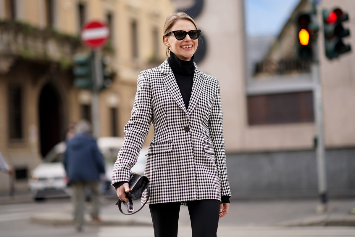 Mulher branca e jovem, de cabelo liso loiro, andando pelas ruas de Paris, na França. Ela usa uma blusa preta de gola alta, óculos escuros, um blazer quadriculado preto e branco, uma calça legging preta e uma bolsa preta, de couro. - Metrópoles