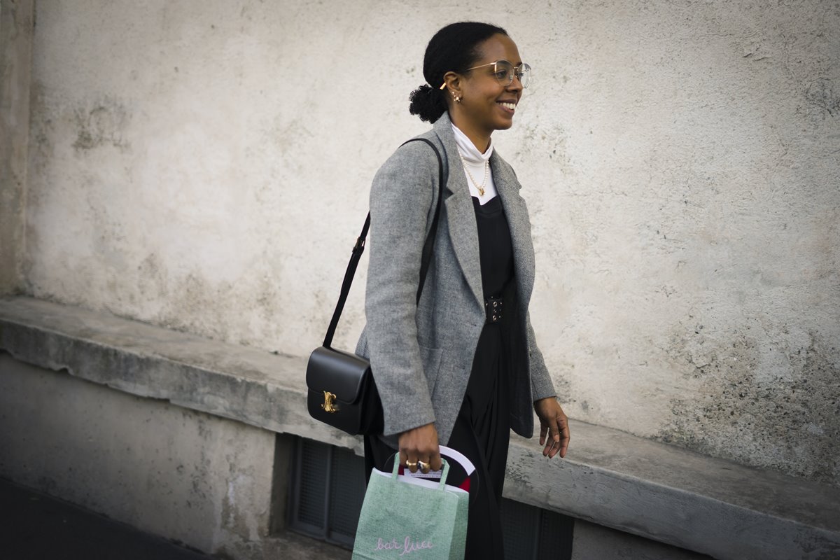 Mulher negra e jovem, de cabelo cacheado curto amarado em um coque, andando pela rua. Ela usa uma blusa de gola alta branca, um vestido preto por cima e um blazer cinza. - Metrópoles