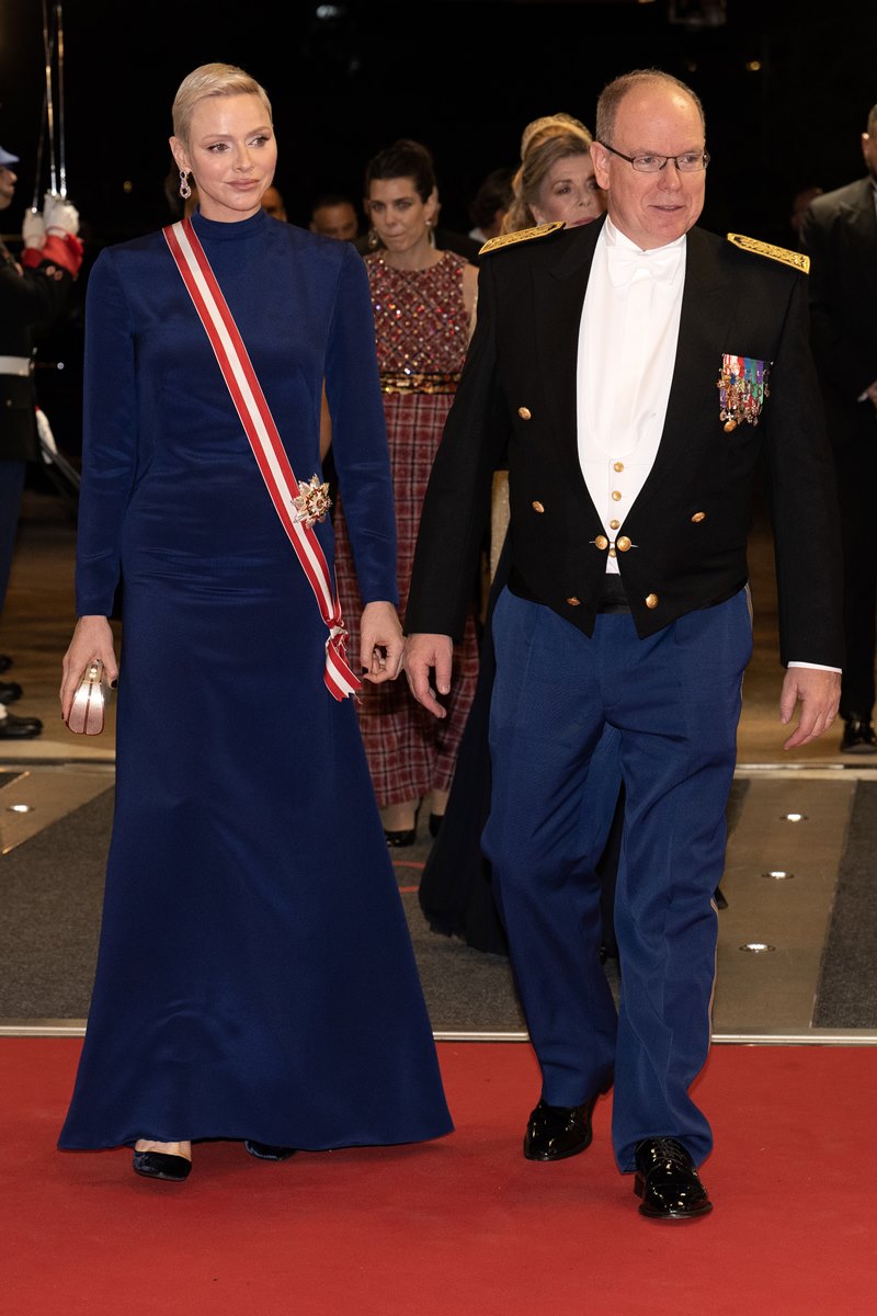 Foto colorida de mulher branca, com cabelo platinado e vestido azul com faixa vermelha ao lado de homem branco, careca e vestido com fraque - Metrópoles