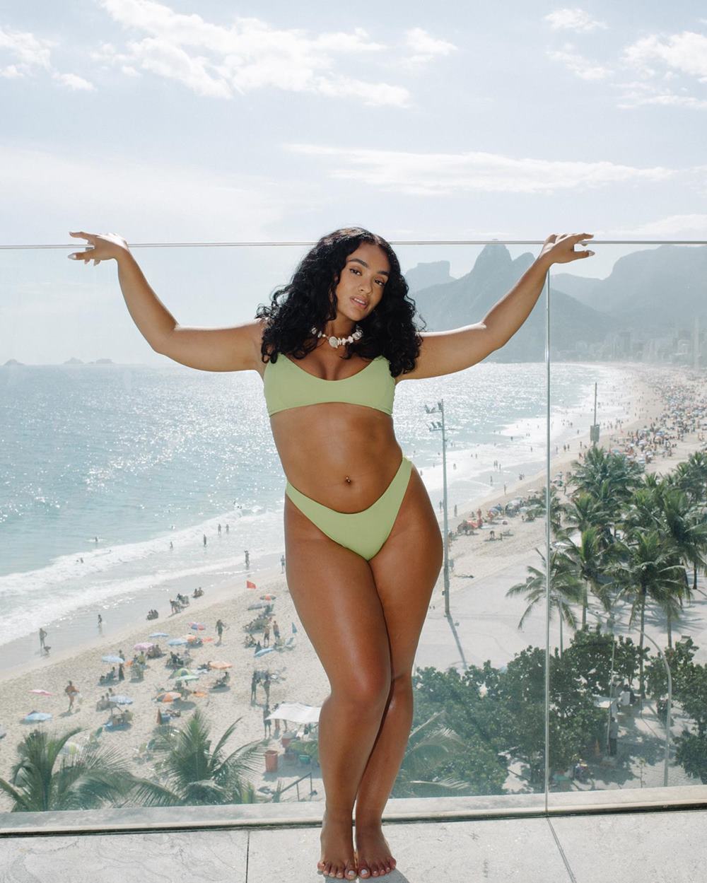 Mulher negra e jovem, de cabelo cacheado preto, posa para foto em um hotel no Rio de Janeiro. No fundo é possível ver a praia e o Pão de Açúcar. Ela usa top e calcinha de biquíni, ambos verde claro. - Metrópoles