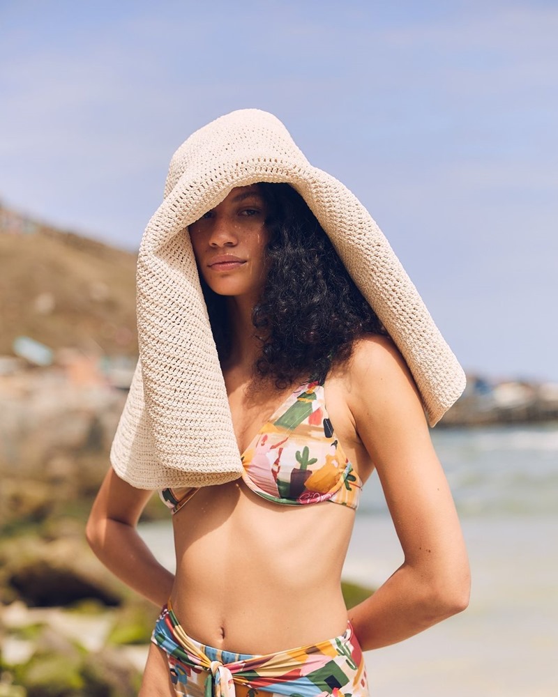 Mulher morena e jovem, de cabelo cacheado médio, posa para foto na praia. Ela usa um biquíni estampado, da marca Ph Praia, e um chapéu de palha. - Metrópoles