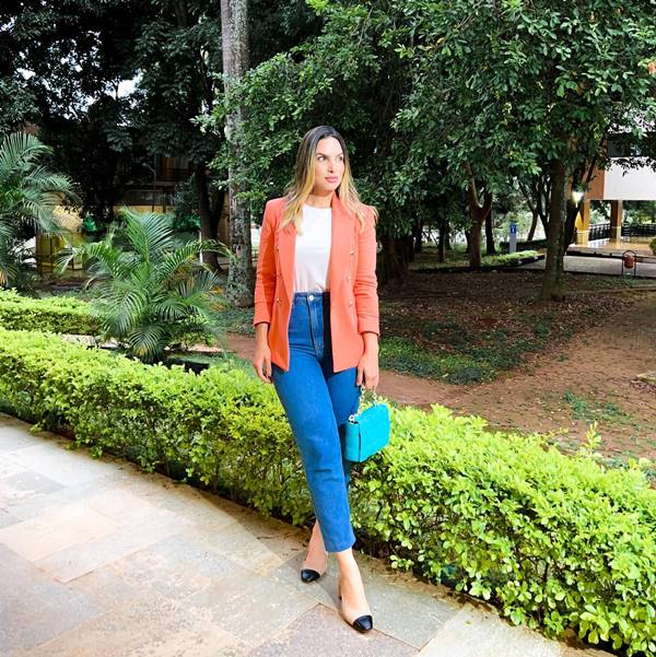 Mulher branca usa blusa branca, blazer laranja e calça jeans - Metrópoles 