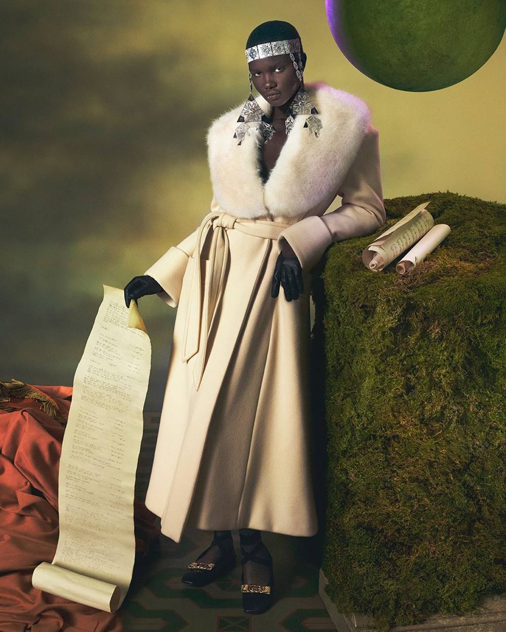 Campanha publicitária da marca Gucci. Na imagem, uma mulher negra e jovem está escorada em uma planta. Ela usa um casaco branco com gola de pelo e joias na cabeça. Ela segura um pergaminho que vai até o chão. - Metrópoles