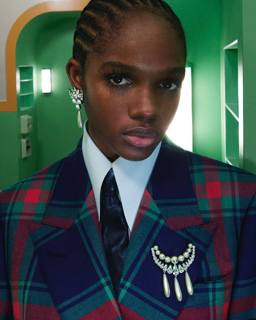 Campanha publicitária da marca Gucci. Na imagem, uma mulher negra e jovem, de tranças, usa camisa branca, gravata e um blazer xadrez, além de um brinco e um brochê de brilhante. - Metrópoles