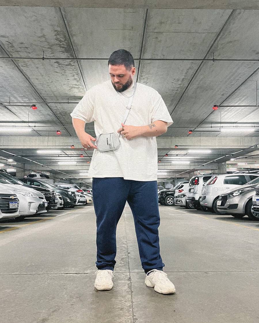 O influencer Vitor Arruda, um homem branco e jovem, de barba preta e cabelo loiro descolorido curto, posa para foto em um estacionamento de carros. Ele usa uma camiseta branca, uma calça preta, um tênis branco Yeezy, da Adidas, e uma bolsa pequena, na cor prata, transpassada no colo. - Metrópoles